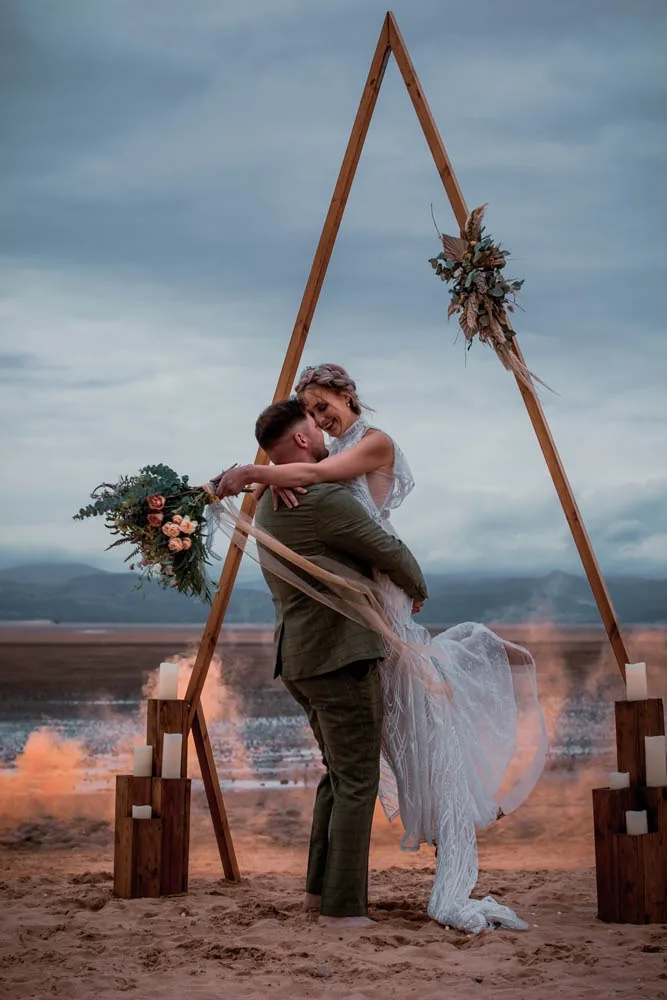 A couple beneath a wooden ceremony arch with the sea behind them
